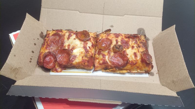 A photo of a Detroit-style pan crust pizza with cheese and pepperoni, on a black table. It's messy but still delicious.